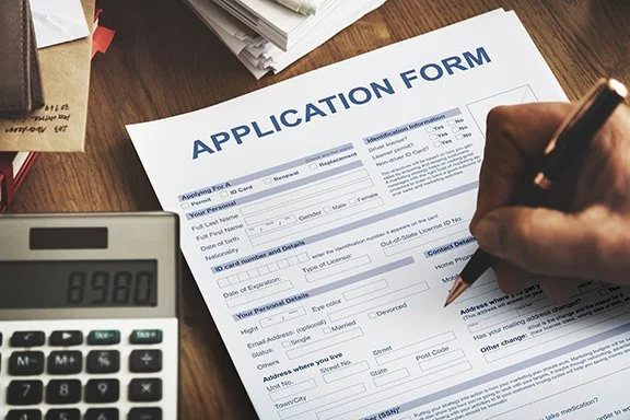 Person filling out an application form on a wooden desk with a calculator nearby.