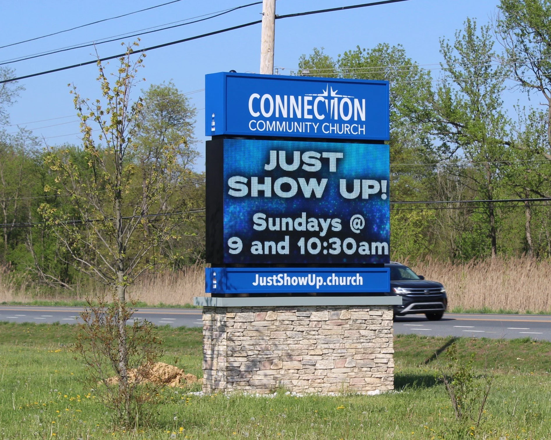 A digital sign outside Connection Community Church announces upcoming Sunday services at 9 and 10:30 am, with a background of trees, power lines, and a passing black vehicle.