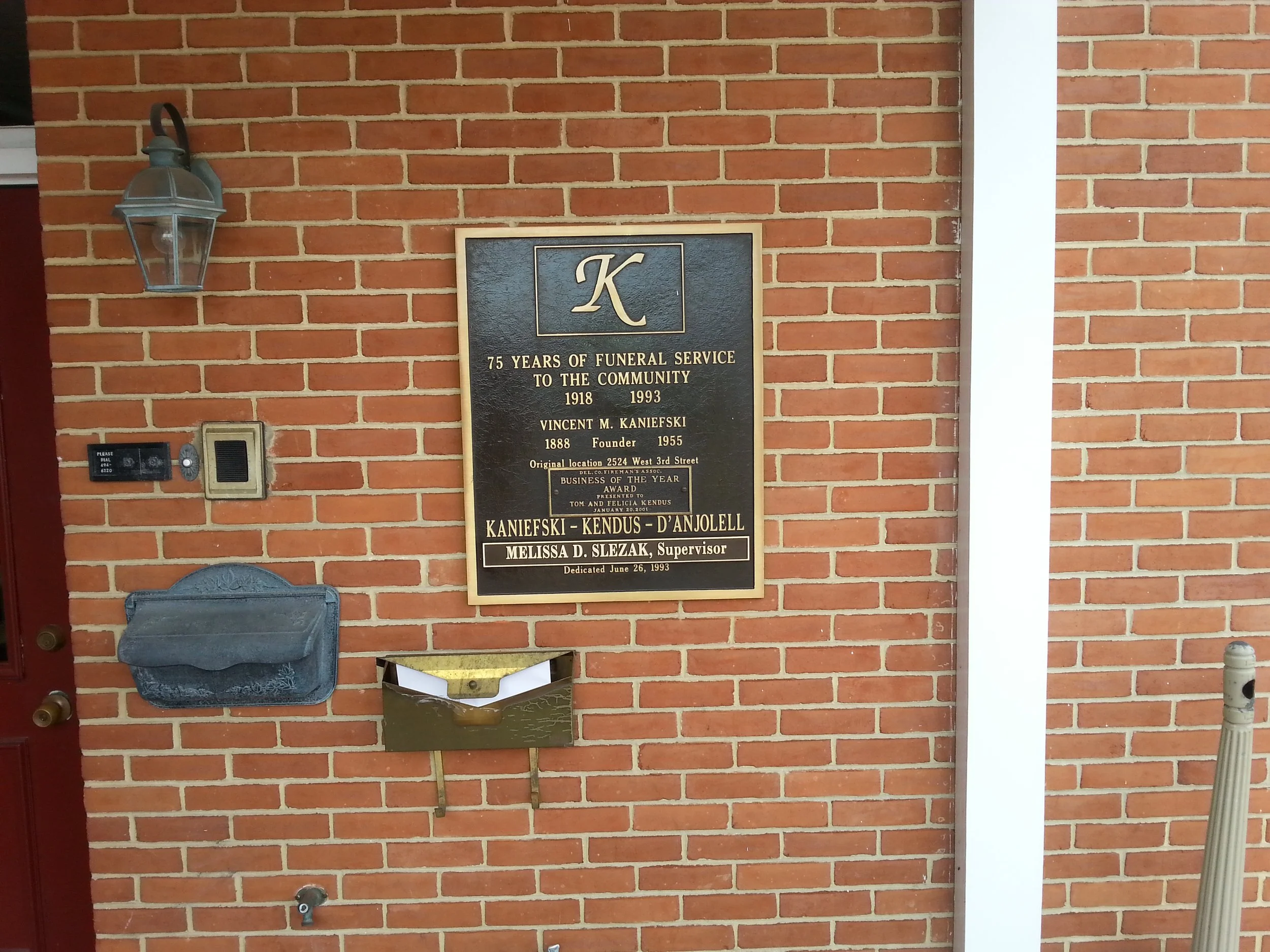Brick wall with a black commemorative plaque celebrating 75 years of funeral service, from 1918 to 1993, in memory of Vincent M. Kaniefski. The plaque also mentions the original location on West 3rd Street, and features the names Kaniefski, Kendus, and D'anjolell, along with Melissa D. Slezak as supervisor. To the left of the plaque, there is an outdoor lantern, a black mailbox, and an intercom system. The lower part of the wall has a metal mailbox and other fixtures.