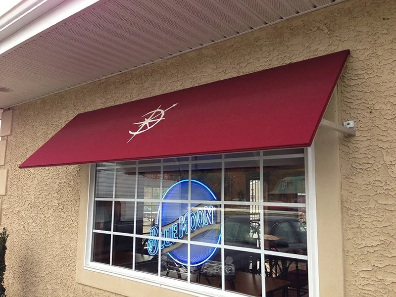 A restaurant or cafe storefront with a red awning and a white logo resembling a compass or nautical instrument. A neon sign inside the window displays the words 'BLUE MOON' in blue.