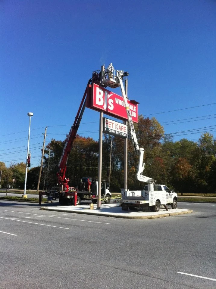 Two utility trucks are working on changing or fixing a sign in a parking lot. One truck has a lift extended to the top of a sign for B's Wholesale, while the other truck is parked nearby.