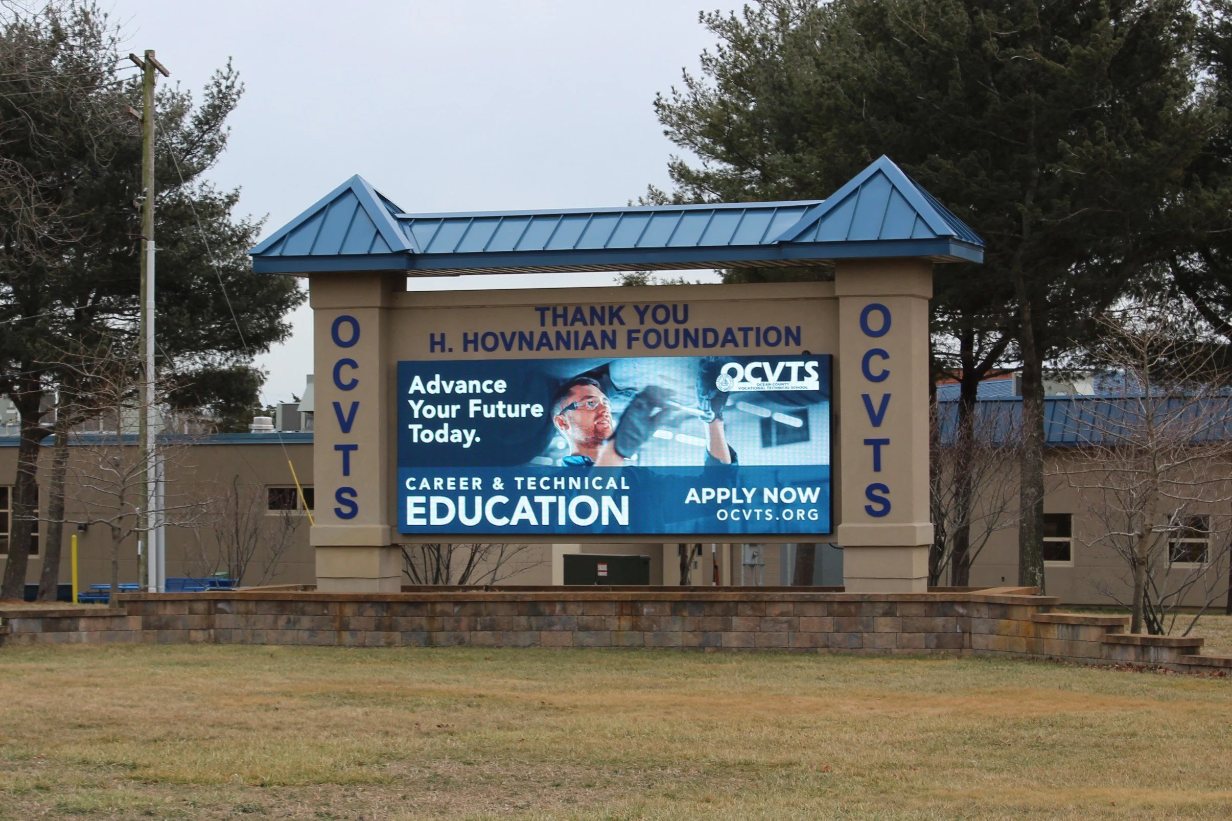 Signboard for H. Hovnian Foundation with a digital display promoting career and technical education, featuring a man wearing glasses and a graduation cap, and the message "Advance Your Future Today. Apply Now".