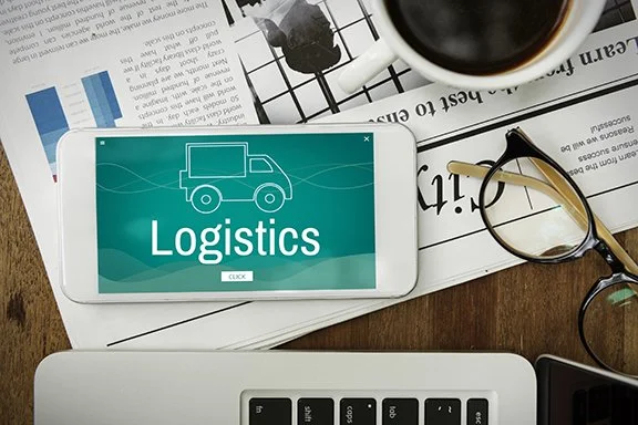 A smartphone displays a logistics icon and text, placed on a cluttered desk with a cup of coffee, reading glasses, a laptop, and some newspapers.