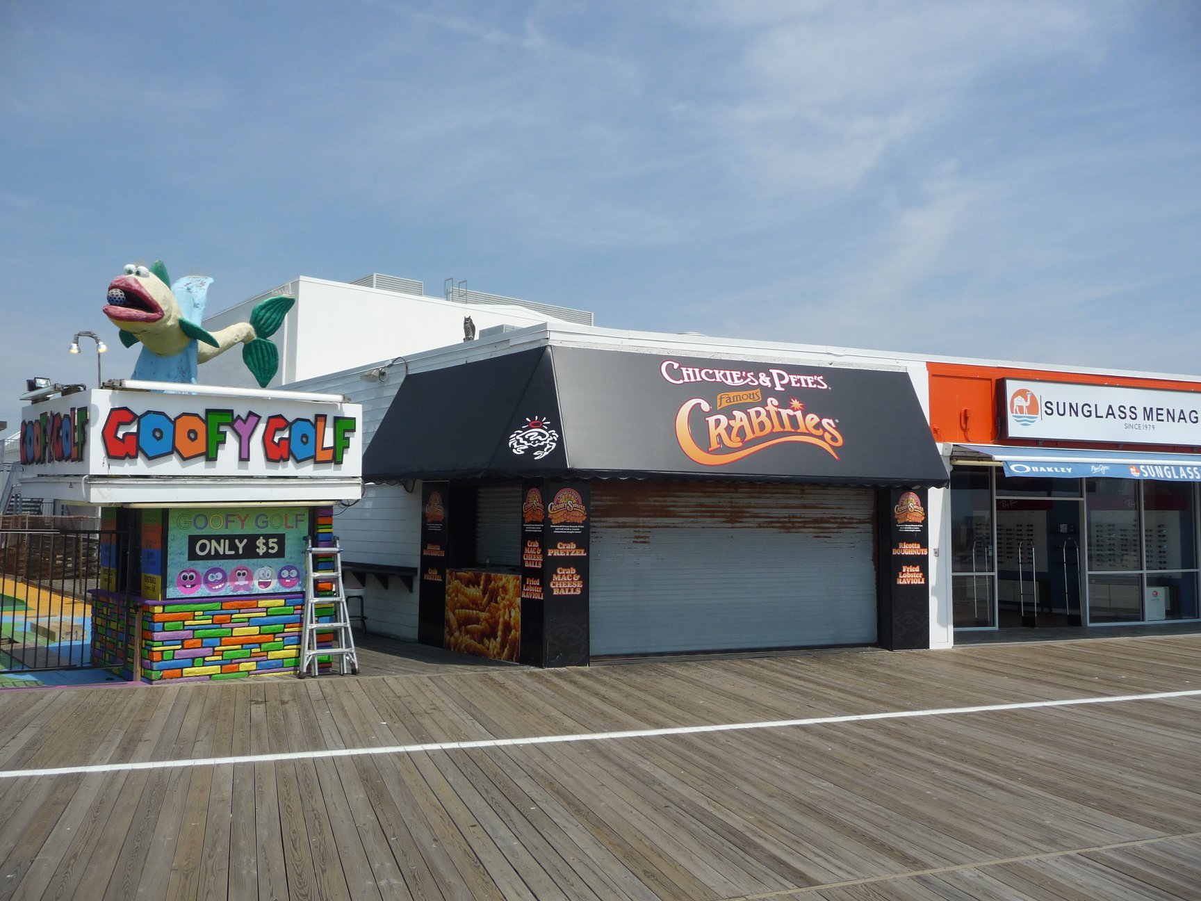 A pier with closed food stands, including Chickie's & Pete's Crab Bites and Sunglass Menag, with a colorful go-kart ride called Gooey Golf and a large fish sculpture on top.