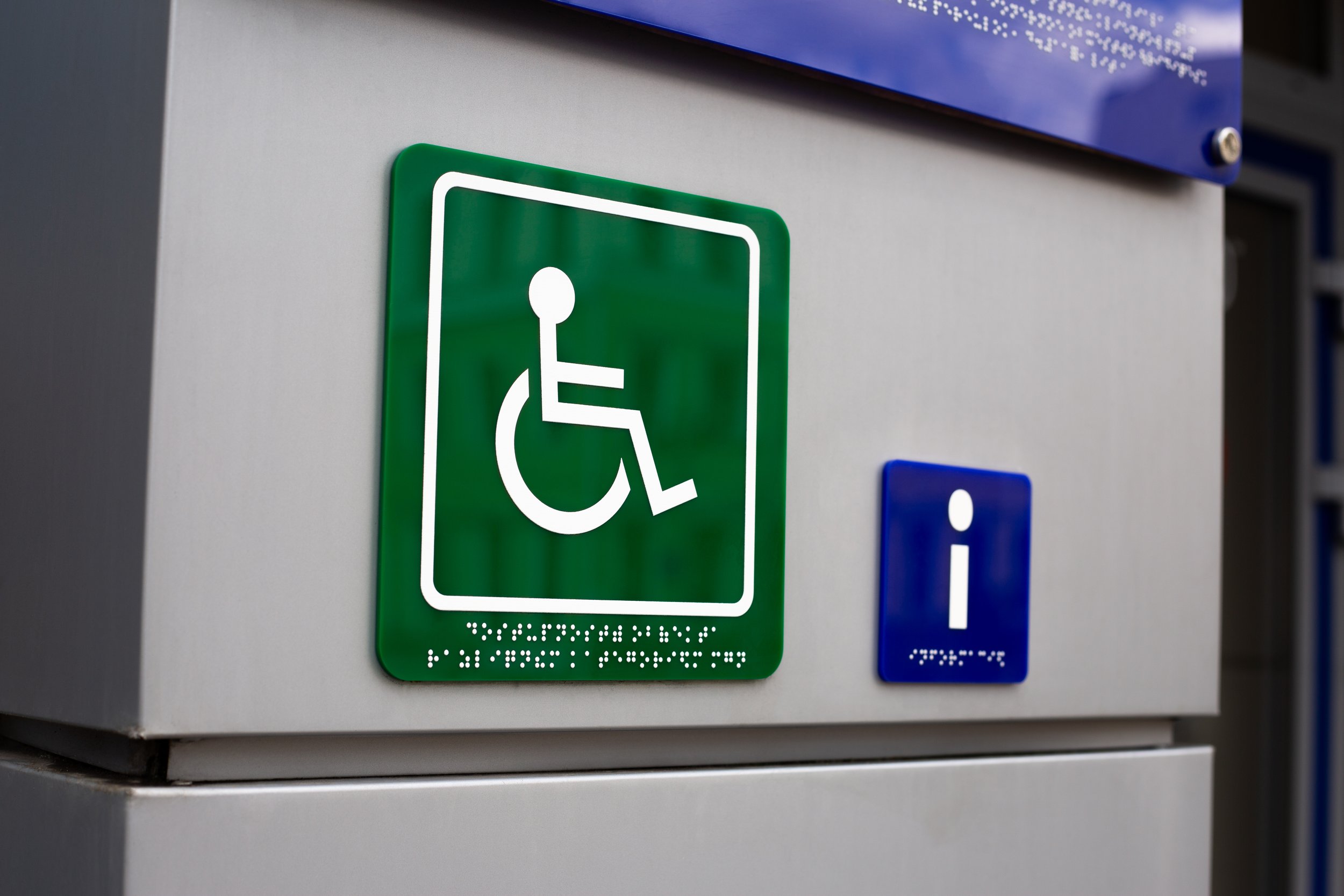 A green accessibility sign with a white wheelchair symbol and Braille text below, mounted on a gray surface, with a blue sign above.