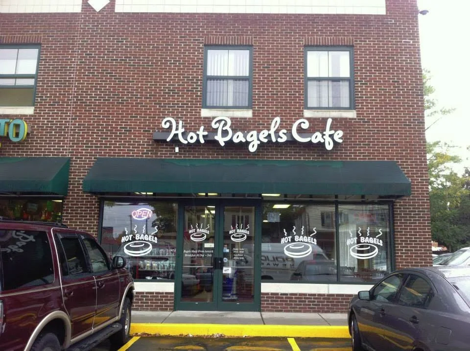 Brick storefront of Hot Bagels Cafe with large windows, green awning, and parking lot in front, featuring a maroon SUV and a dark gray car.