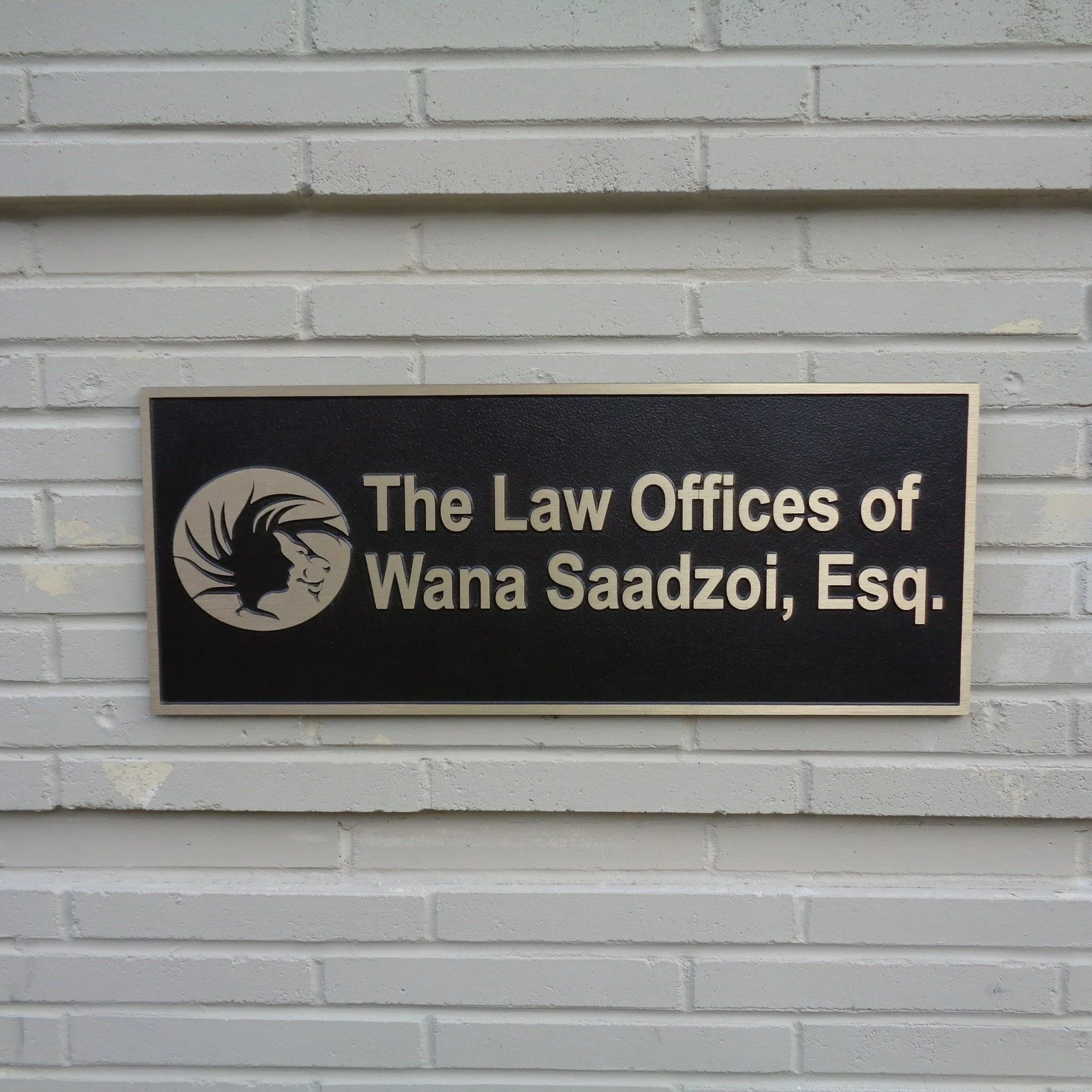 Sign on a white brick wall that reads 'The Law Offices of Wana Saadzo, Esq.' with a logo of a woman's profile inside a circle.