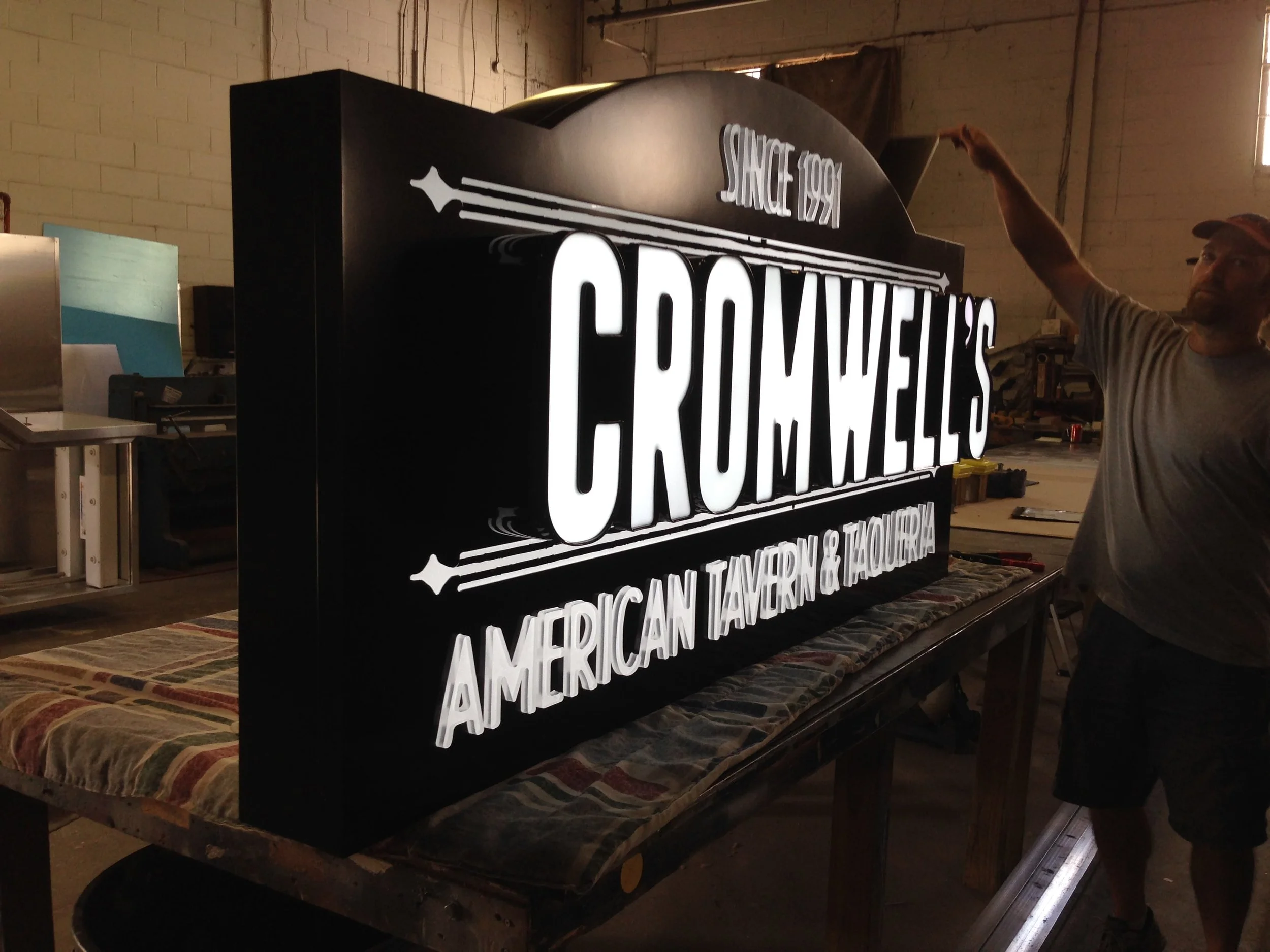 A person in a gray shirt and cap is adjusting or installing a large illuminated sign that reads 'KITCHEN 1992' and 'FRENCH, ITALIAN, TAVERN & TACOS.' The sign is on a table inside a workshop with tools, a metal box, and a striped cloth cover.