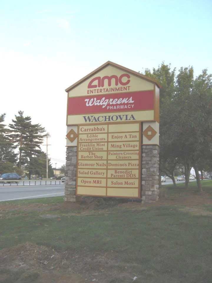 A shopping center sign with multiple storefront signs, including AMC Entertainment, Walgreens Pharmacy, Wachovia, Carrabba's, and various local businesses like a nail salon, pizza place, dental office, and a salon.