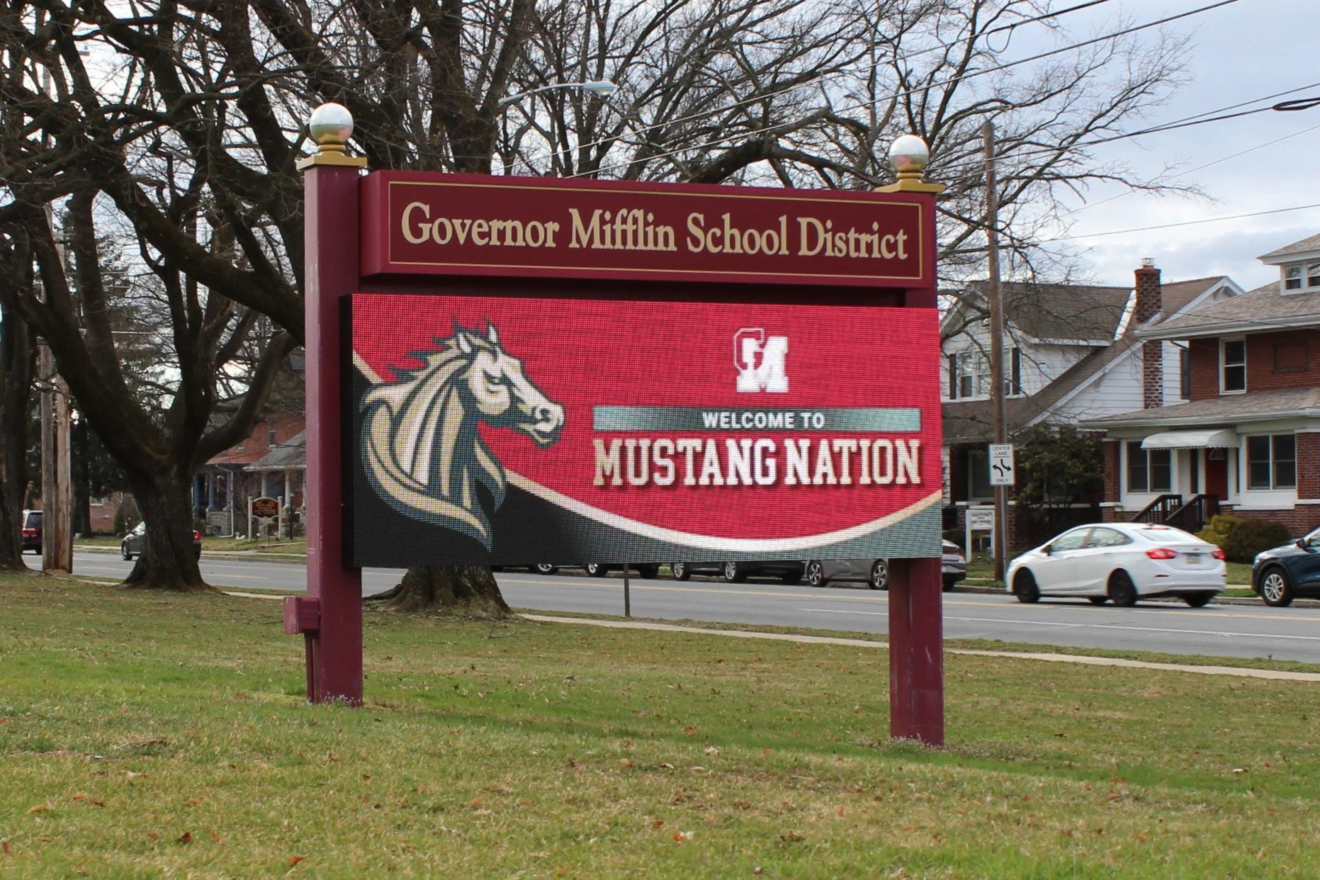 A digital sign on a maroon post displaying the message 'Welcome to Mustang Nation' with a stylized horse head logo, situated in front of trees and residential houses along a street with cars passing by.