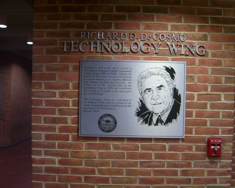 Plaque dedicated to Richard D. DeCosmo with a portrait illustration on a brick wall in the Technology Wing.