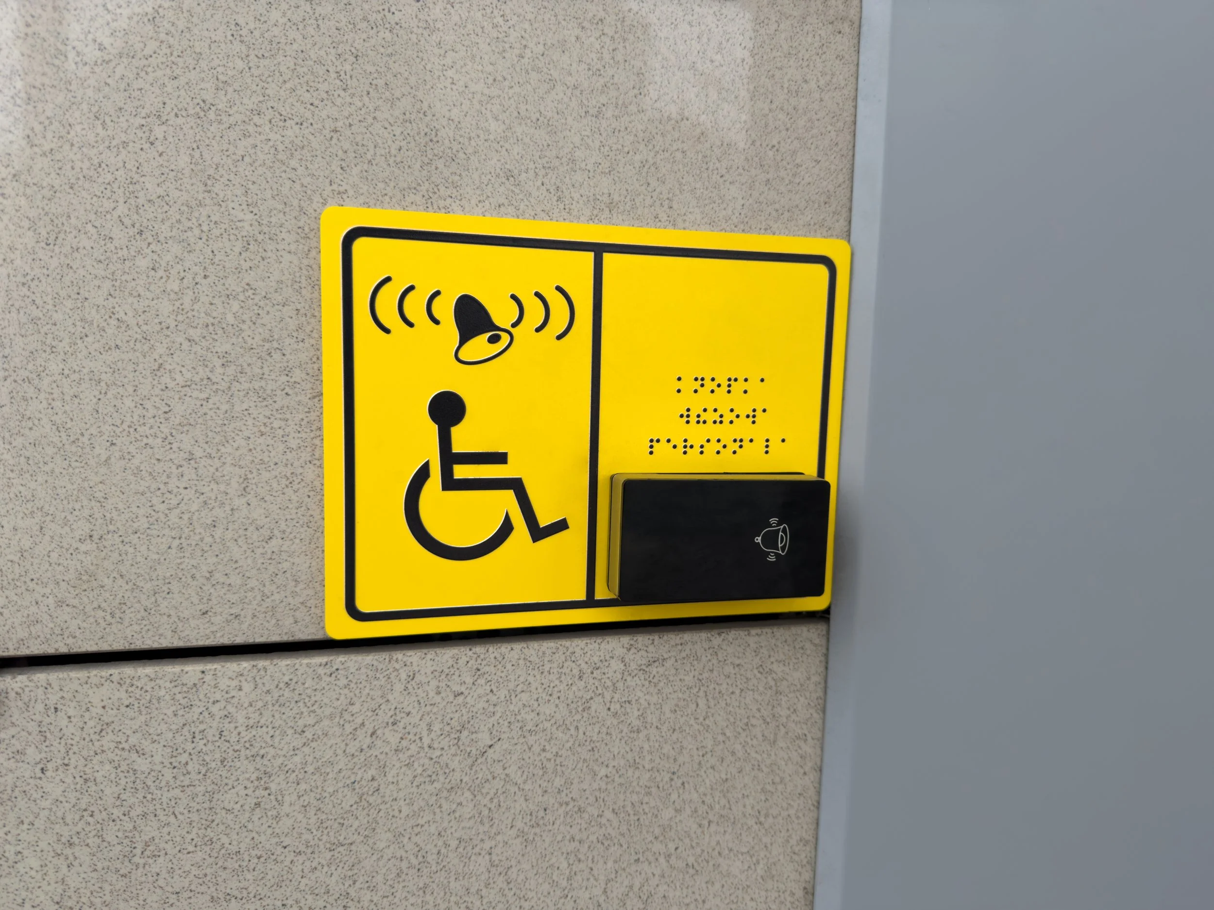 Yellow accessibility sign with a wheelchair symbol and a bell icon, mounted on a wall next to a gray door.