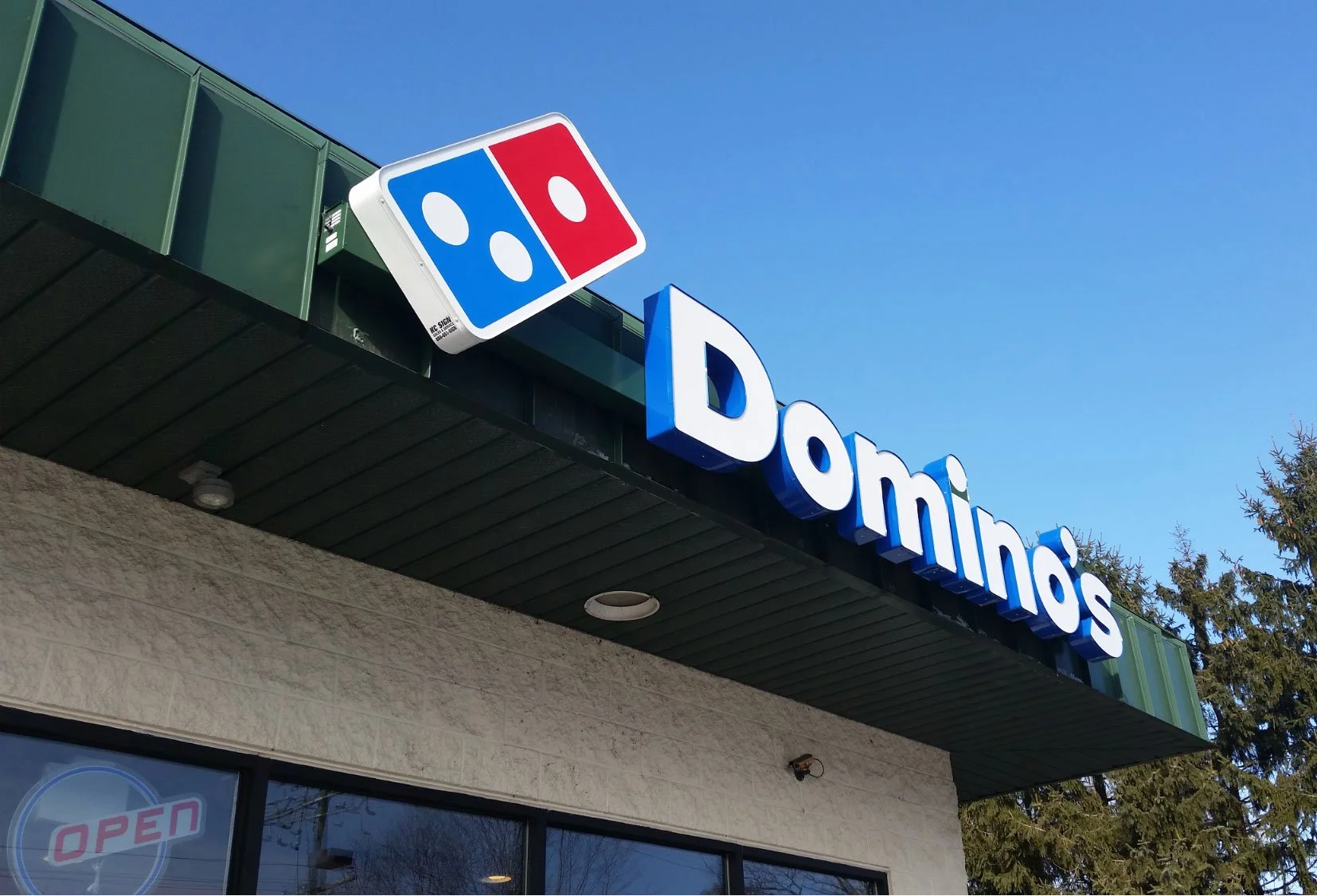 Exterior of a Domino's Pizza restaurant with a sign showing the Domino's logo and the name 'Domino's' in blue letters underneath; an 'Open' sign can be seen in the window.