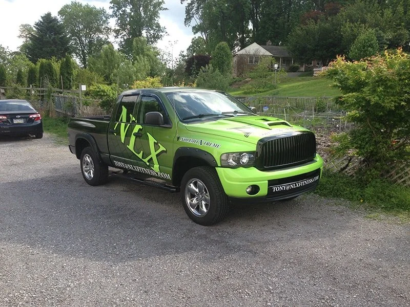A lime green and black pickup truck with large NEX lettering on the side, parked on a gravel lot next to a black sedan. The truck has a website URL on the bumper and is near trees and a fence.