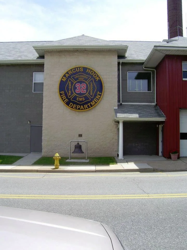 Building with a circular sign that reads 'Marcus Hook Fire Department' with the number 32 in the center, and the words 'Organized 2003' and 'EMS' around it. A fire hydrant and a bell on the sidewalk in front.