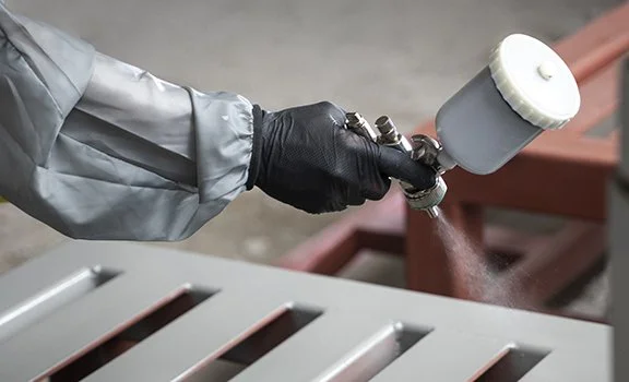 A person wearing a gray jacket and black gloves holding a spray gun and applying a coating to metal slats outdoors.