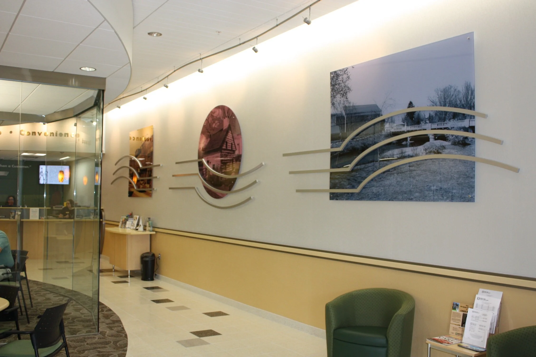 Interior of a public building, likely a waiting area, with chairs, wall art, and a reception desk. The wall features landscape photographs, and there are ceiling lights and a curved design element.
