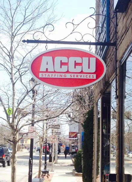 Outdoor store sign for ACCU Staffing Services hanging from decorative metal bracket on building.