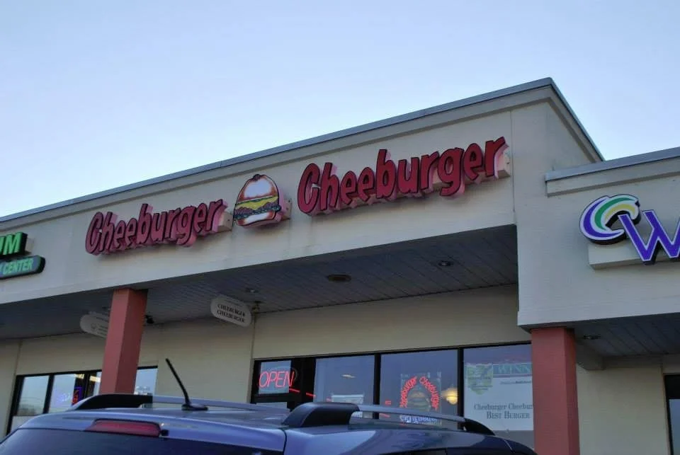 The storefront of a Cheeburger Cheeburger fast food restaurant with a pink sign, a burger logo, and an illuminated 'Open' sign in the window.