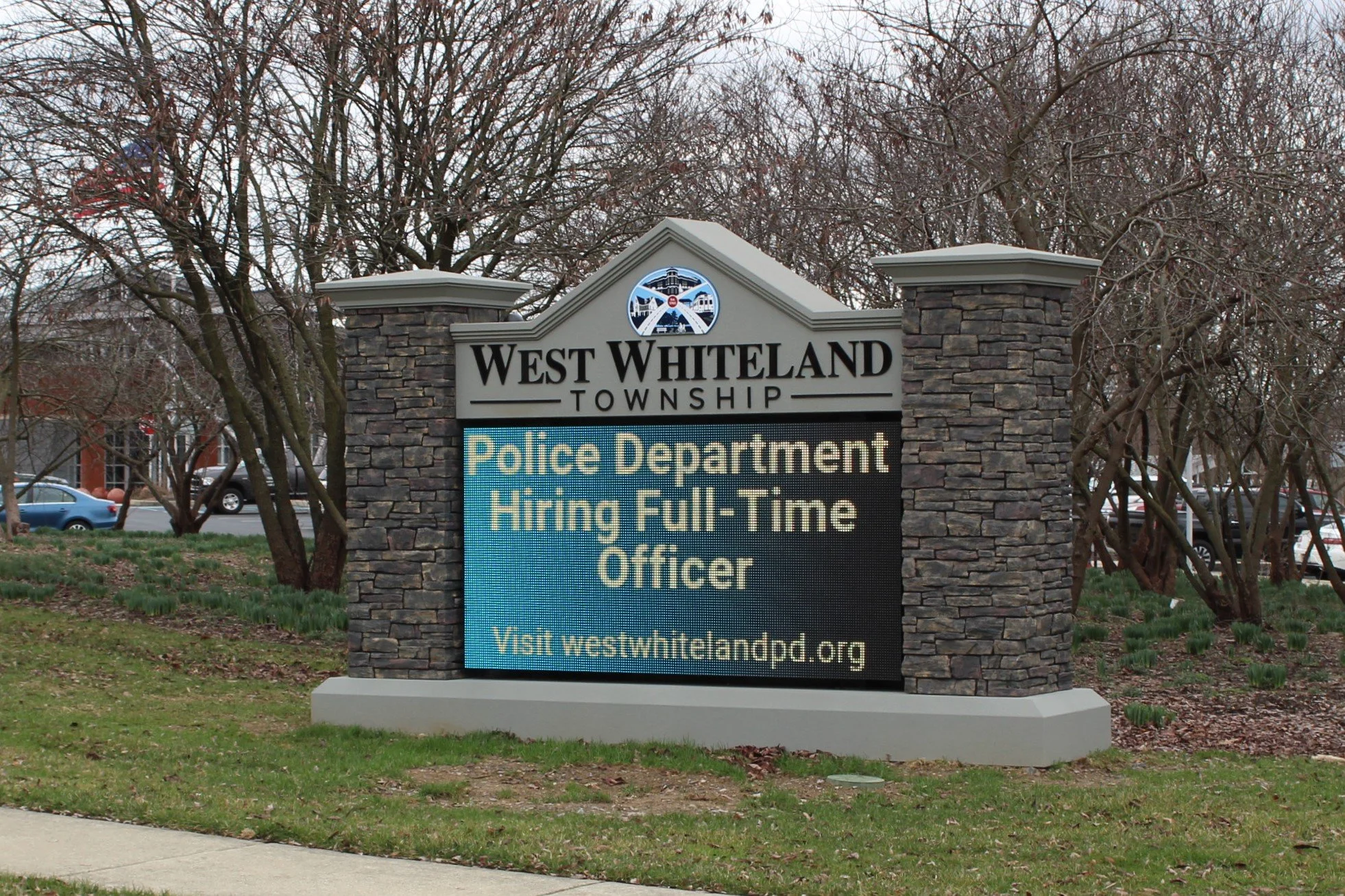 A sign for West Whiteland Township Police Department announces job opening for a full-time officer, with a website link www.westwhitelandpd.org. The sign is placed outdoors with trees and cars visible in the background.