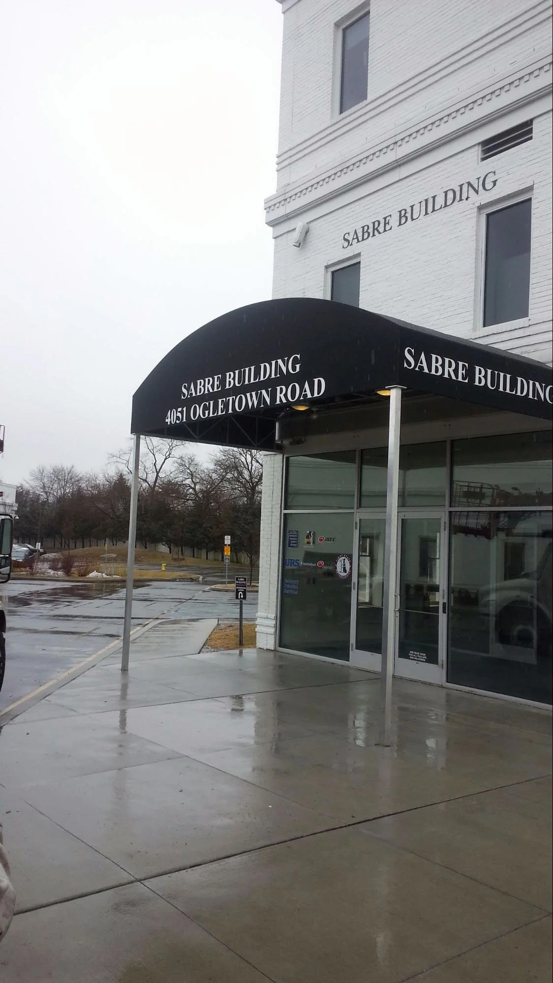The image shows the entrance of the Sabre Building, with a black awning that has the address 4051 Ogle T own Road and the building name printed on it. The building is a white brick structure with several windows, and the entrance has glass doors. The