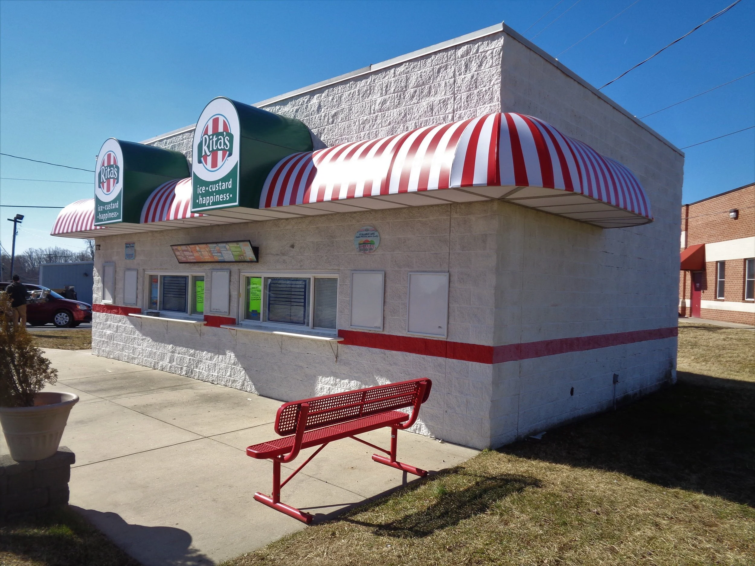 A small fast food restaurant with a red and white striped awning, green signs with the Rita's logo, and a red outdoor bench in front. There are large windows on the building and a parking lot with cars behind it.