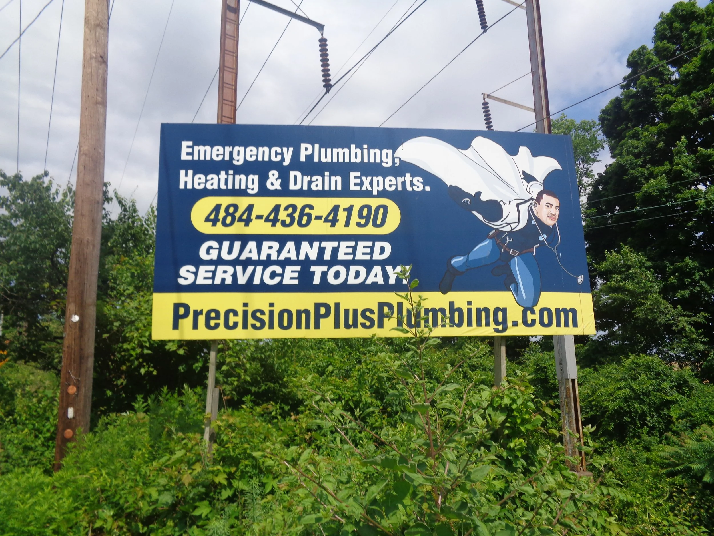 Blue sign advertising emergency plumbing, heating, and drain services, with a cartoon mascot of a plumber holding a wrench, and the phone number 484-436-4190. The sign is outdoors with green foliage and utility poles in the background.
