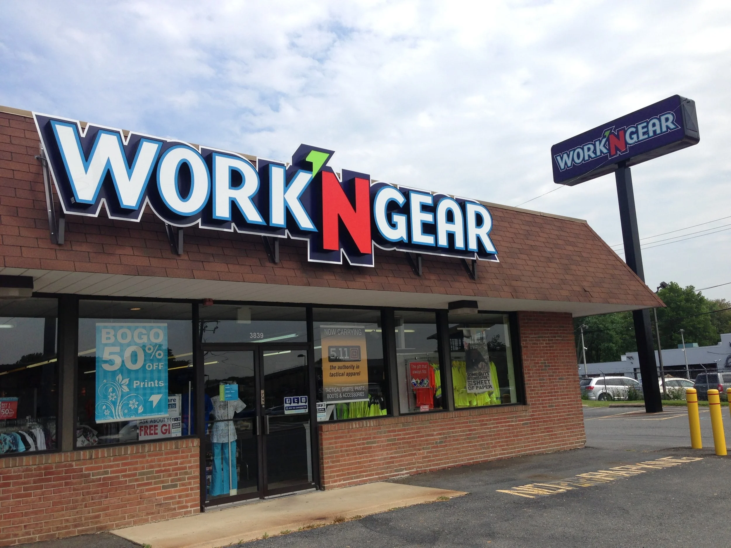 A retail store named 'Work'n Gear' with a large sign on the building's exterior and a smaller sign on a tall pole, with visible storefront windows displaying clothing and signs advertising sales and products.