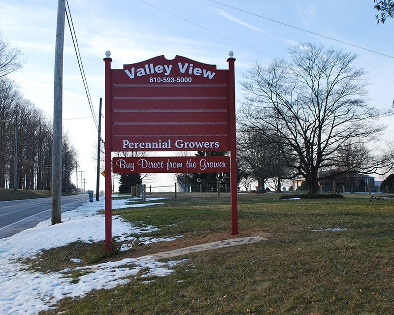 A red and white Valley View sign advertising perennial growers and direct purchase from the grower, placed on a grassy area next to a road with snow patches, trees, and houses in the background.