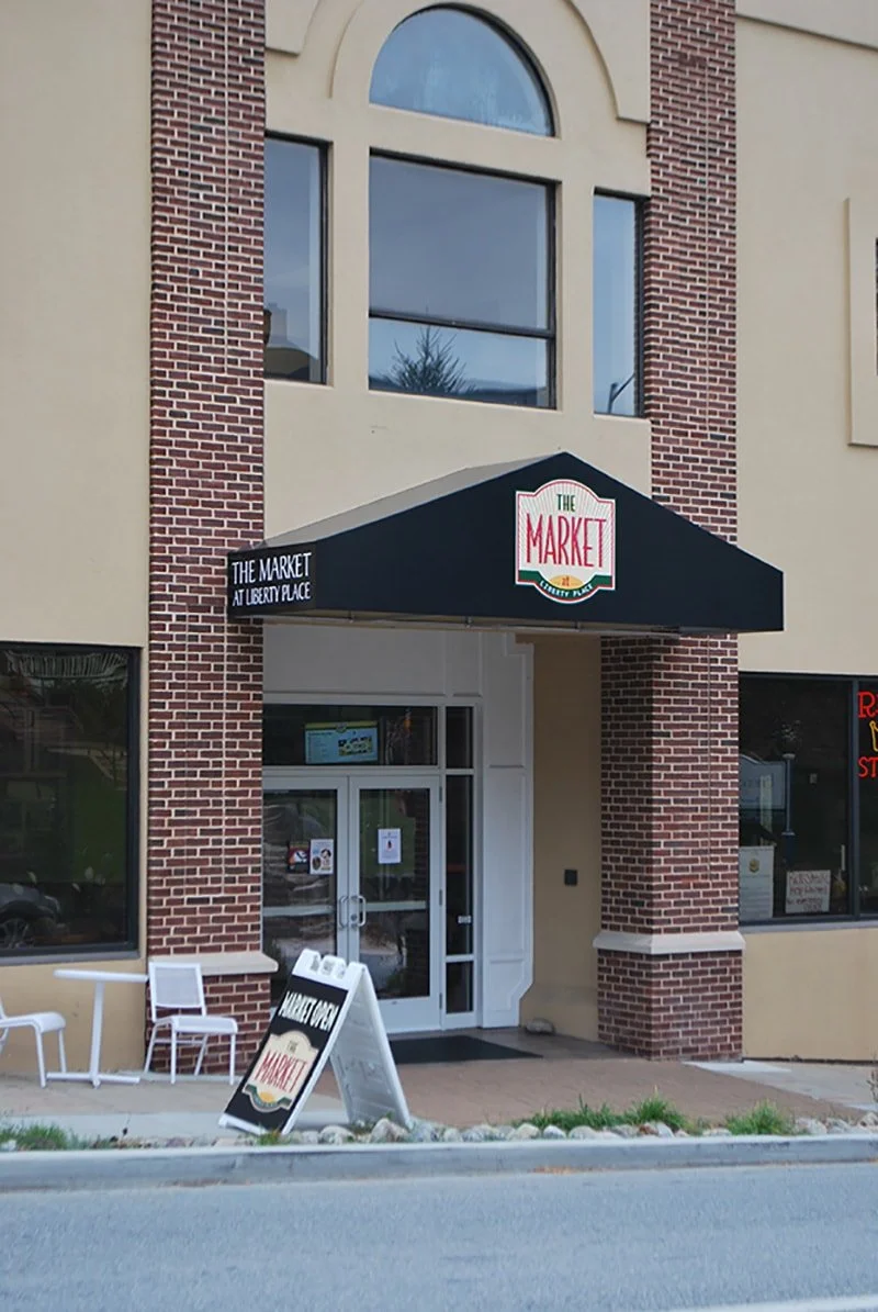 Exterior of a building with a sign reading "The Market" above the entrance and a sandwich board sign outside. The building has brick and beige walls, with large windows.