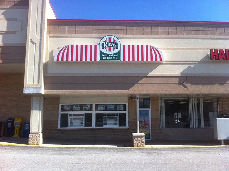 Front view of a Rita's Ice store with a red and white striped canopy, a sign with Rita's logo, and promotional posters in the window.