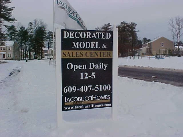 Sign advertising a decorated model and sales center for Jacobucci Homes, open daily 12 to 5, in a snowy landscape with trees and houses in the background.