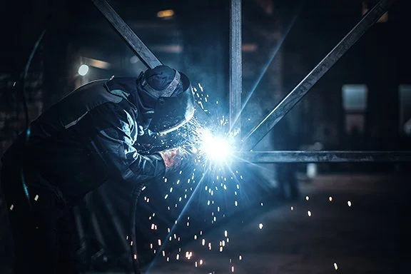 Welder wearing protective gear welding metal beams with sparks flying in a workshop.