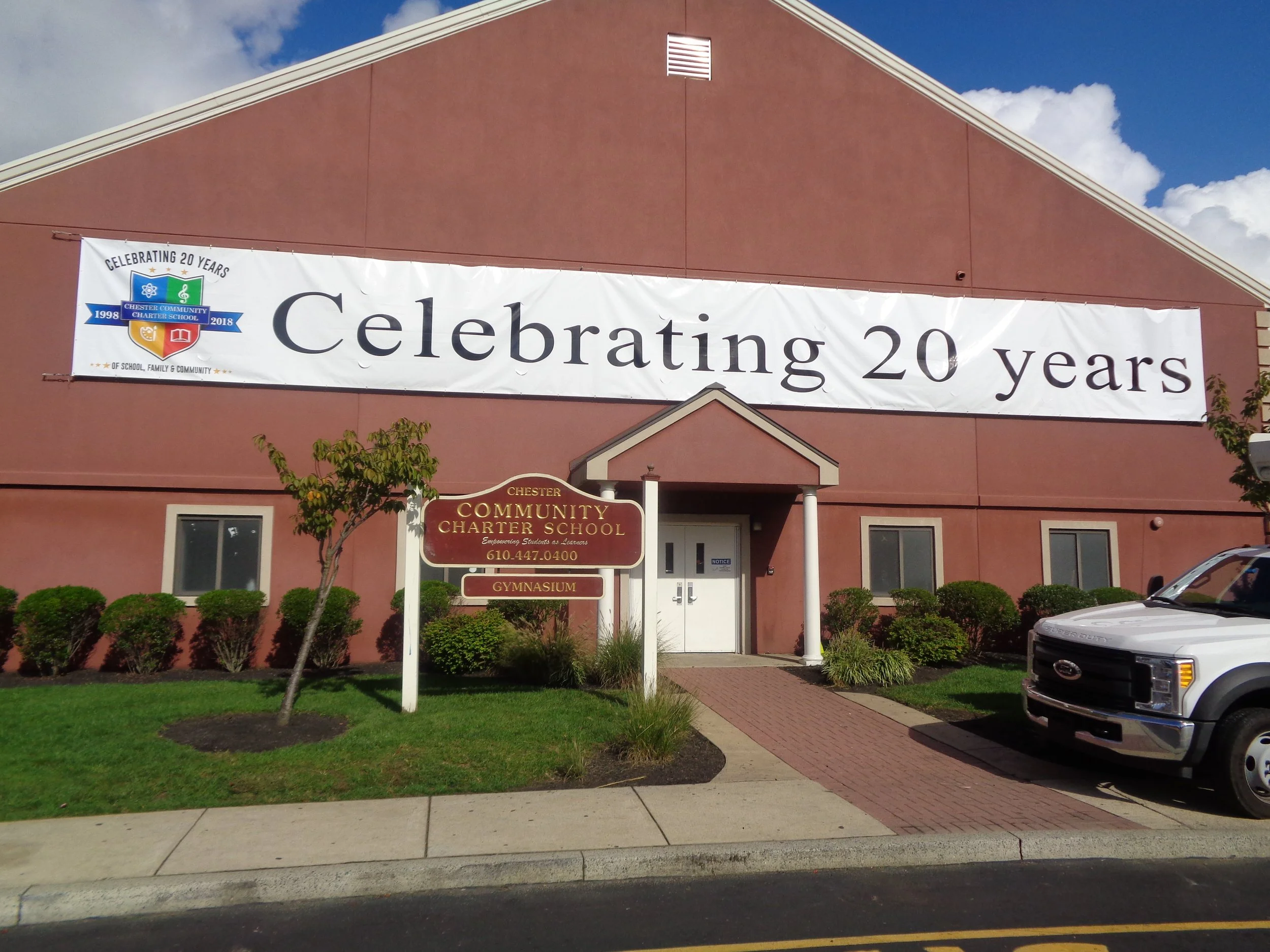 The exterior of a building with a large banner celebrating 20 years of Chester Community Charter School, with trees, a sidewalk, parked cars, and a blue sky with clouds in the background.