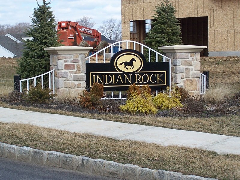 Welcome sign for Indian Rock community with a black background, gold lettering, a silhouette of a horse above the text, stone pillars, and landscaping with shrubs and pine trees.