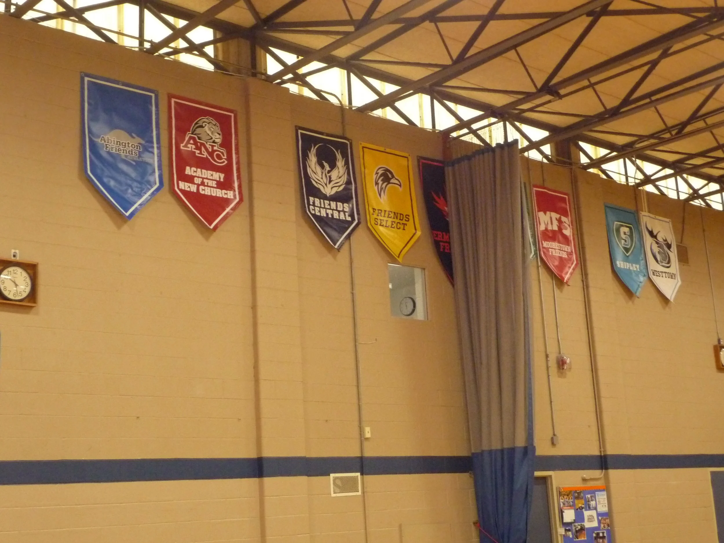 Indoor gymnasium with banners hanging from the ceiling, a clock on the wall, and a display board in the corner.