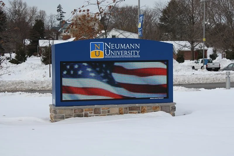 Digital signboard at Neumann University displaying an American flag in a snowy outdoor setting.