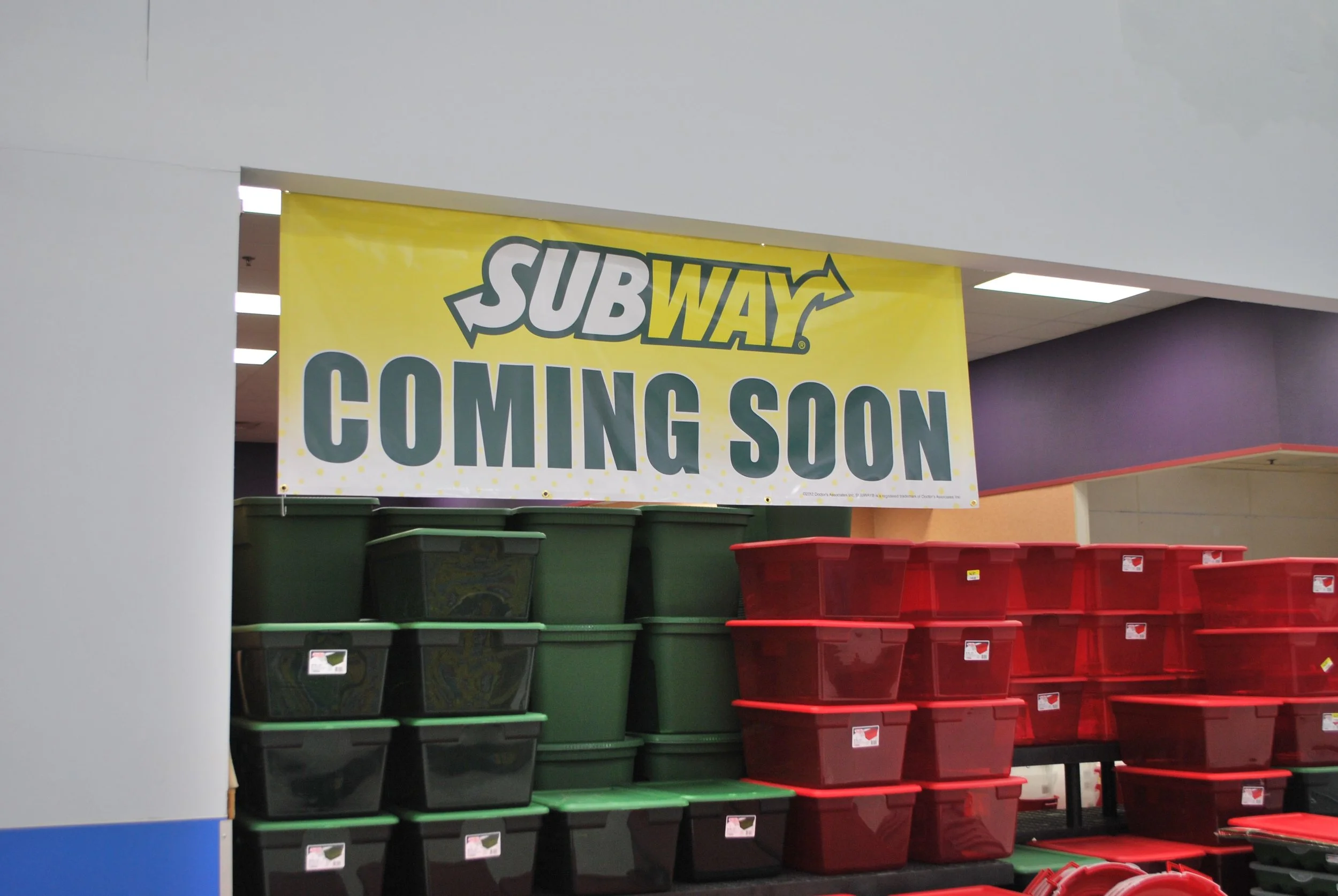 A yellow Subway banner with the words 'Coming Soon' hangs above stacked plastic storage bins in red and green, inside a shopping mall or similar indoor retail space.