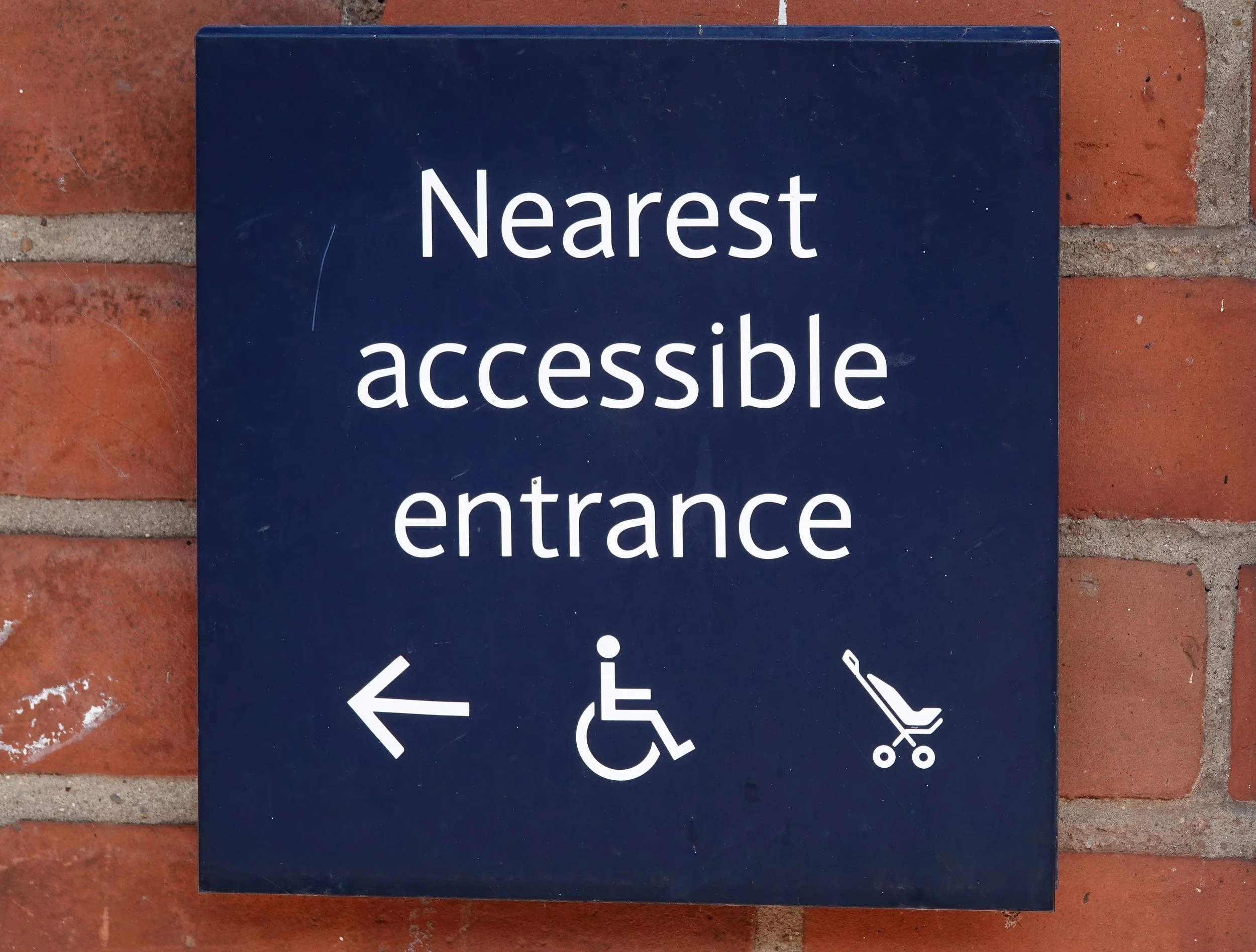 Sign indicating accessible entrance with arrows pointing to the left and straight ahead, and symbols for wheelchair accessible and stroller accessible pathways, mounted on a brick wall.