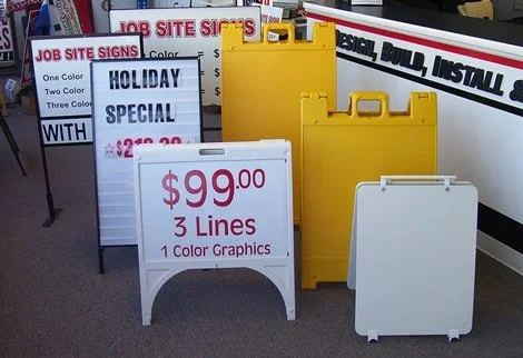 Various portable sandwich board signs, some yellow and white, display prices and advertising text in a store aisle.