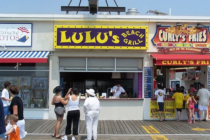 People standing in front of Lulu's Beach Grill and Curly's Fries food stands at a Boardwalk, with seating and menu signs visible.