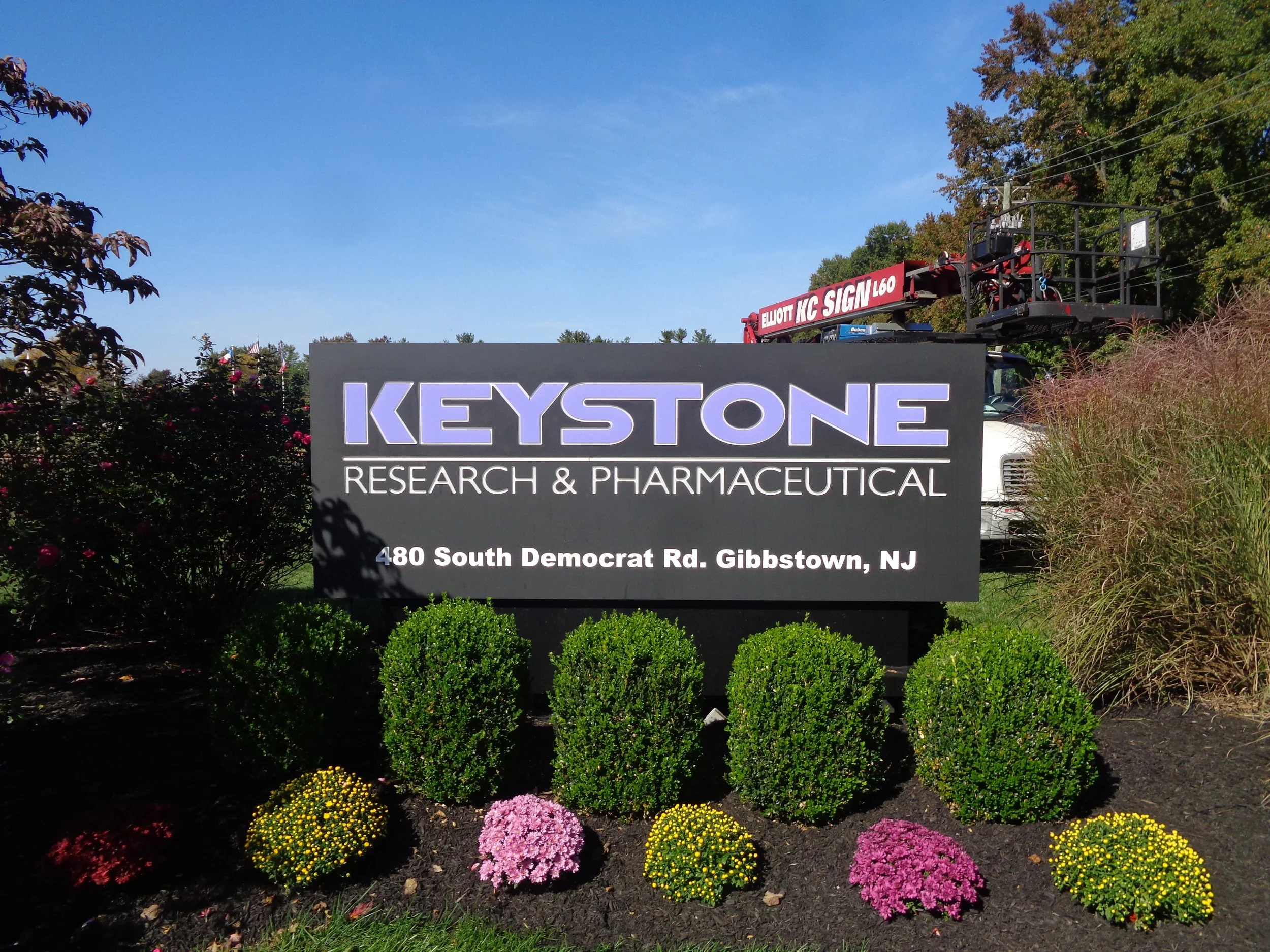 Sign for Keystone Research & Pharmaceutical with flowering plants in front and construction equipment in the background.