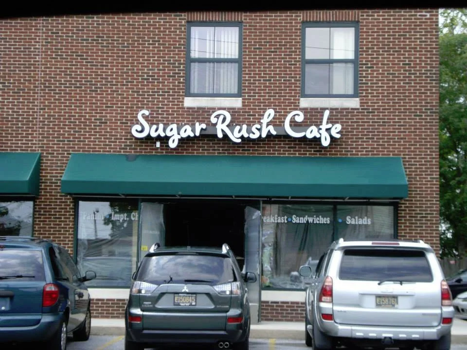 Restaurant called Sugar Rush Cafe with cars parked in front, green awning, and brick building