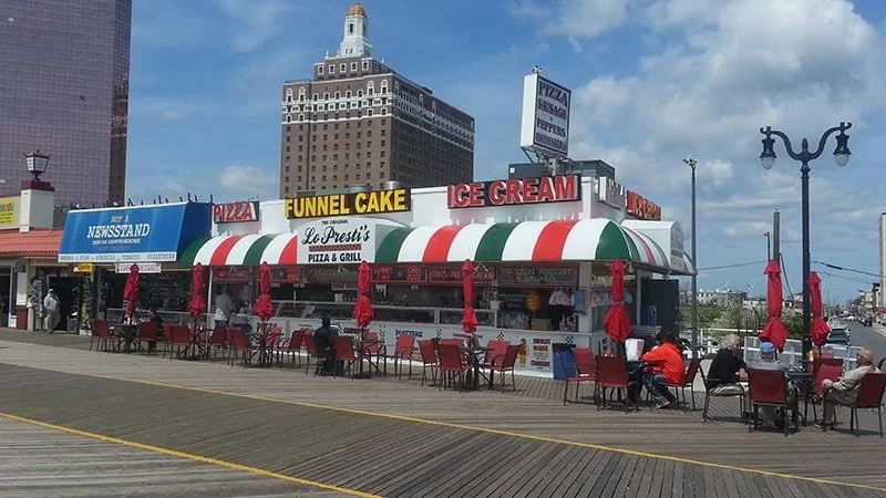 Pier with outdoor seating, red umbrellas, and a food stand selling pizza, funnel cake, and ice cream, with a city skyline in the background.