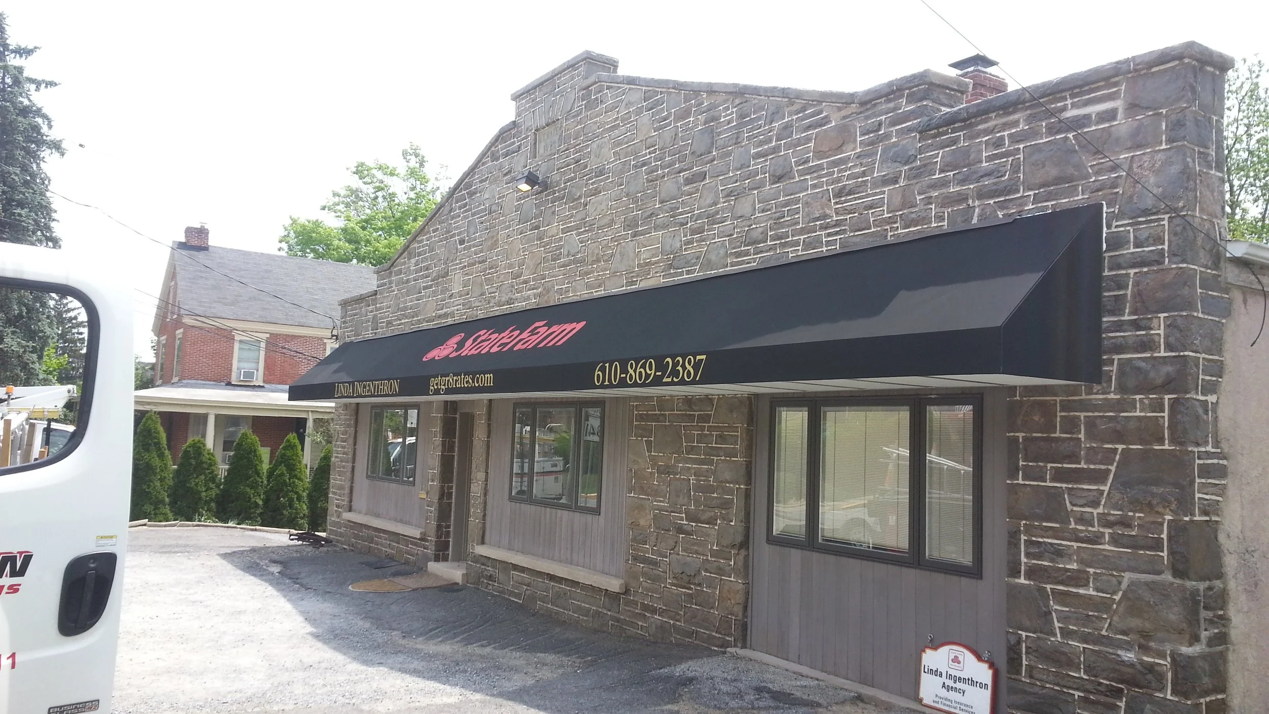 The storefront of a business with a black awning that has pink text reading 'StateFarm' and yellow text with a phone number and website. The building has stone exterior walls and large windows.