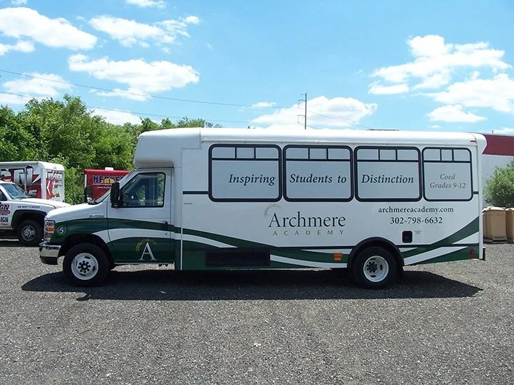 A white and green school bus with advertising for Archmere Academy, highlighting inspiring students to distinction for grades 9-12, parked outdoors on gravel under a blue sky with clouds.