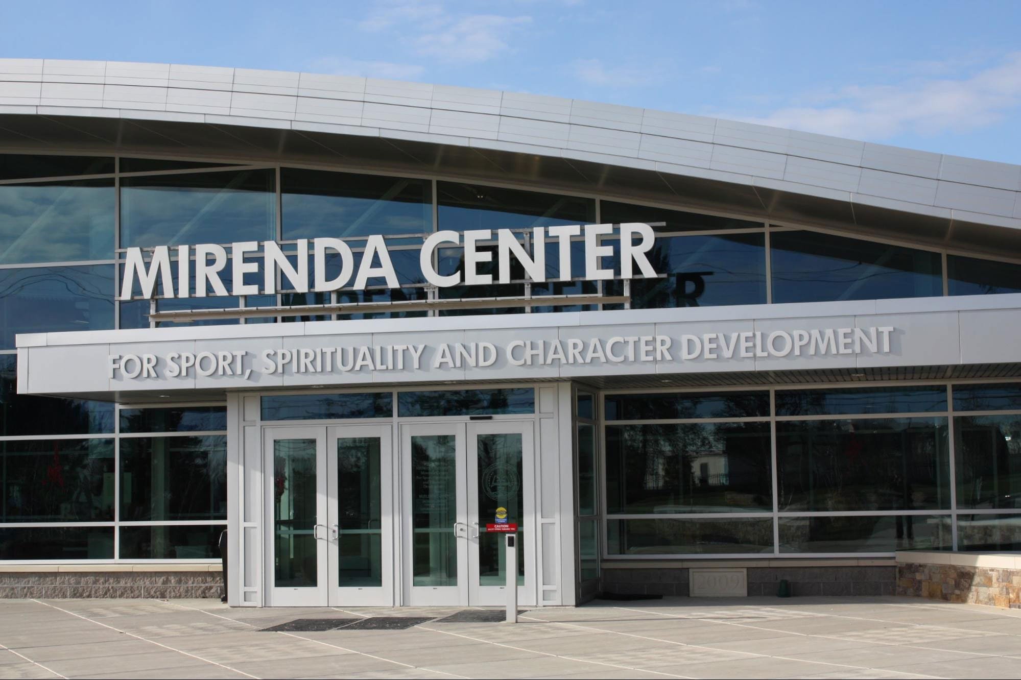 Exterior of the Mirenda Center building with glass windows and doors, featuring prominent white signage that reads "MIRENDA CENTER" and a smaller sign below that says "FOR SPORT, SPIRITUALITY AND CHARACTER DEVELOPMENT."