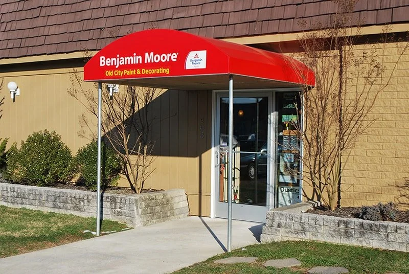 Exterior view of a store named Benjamin Moore with a red awning that reads 'Old City Paint & Decorating.' The entrance has glass doors and is flanked by small garden beds with bushes and trees.