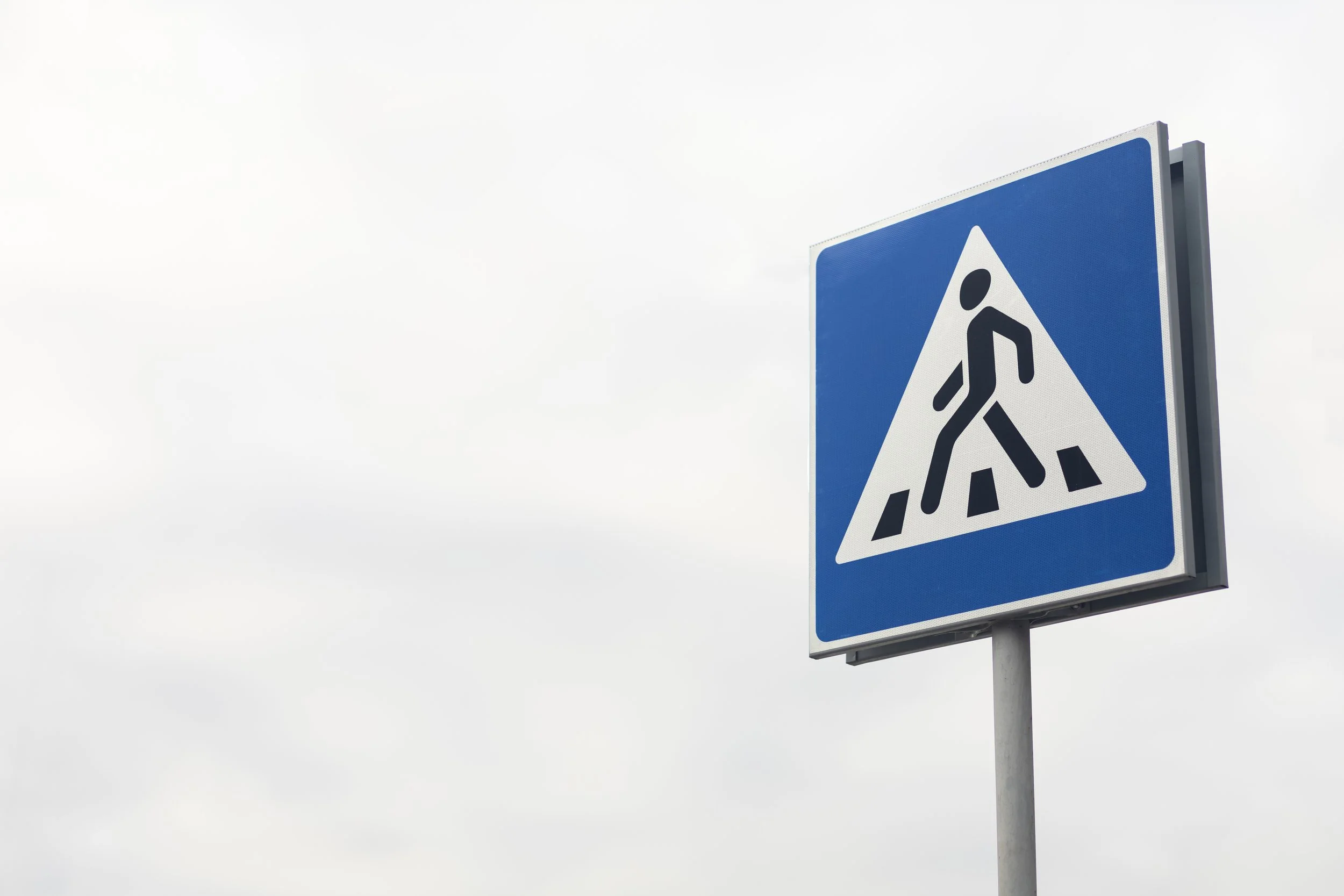 A blue traffic sign with a pedestrian walking on a crosswalk icon, posted against an overcast sky.