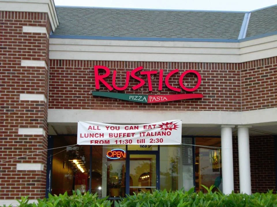 Brick restaurant named Rustico that offers pizza and pasta. A sign advertises all-you-can-eat lunch buffet Italian style for $6.99 from 11:30 AM to 2:30 PM. There is an illuminated 'Open' sign and the entrance features glass doors.